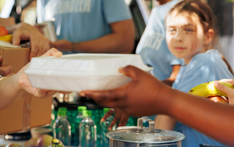 A hot meal container passed from one pair of hands to another at a food distribution